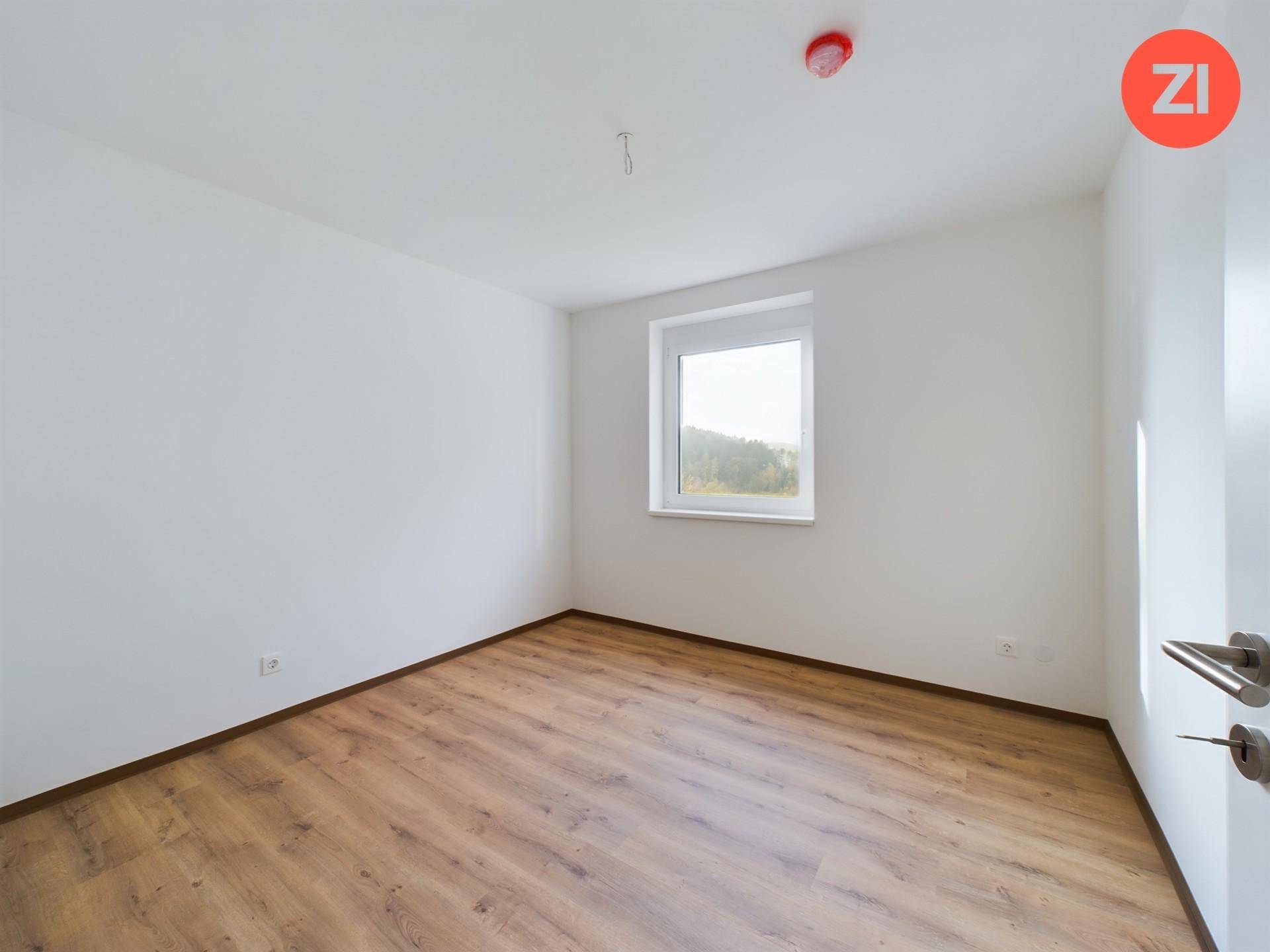 Leeres Schlafzimmer mit Laminatboden, weißen Wänden und einem Fenster mit Blick ins Grüne.