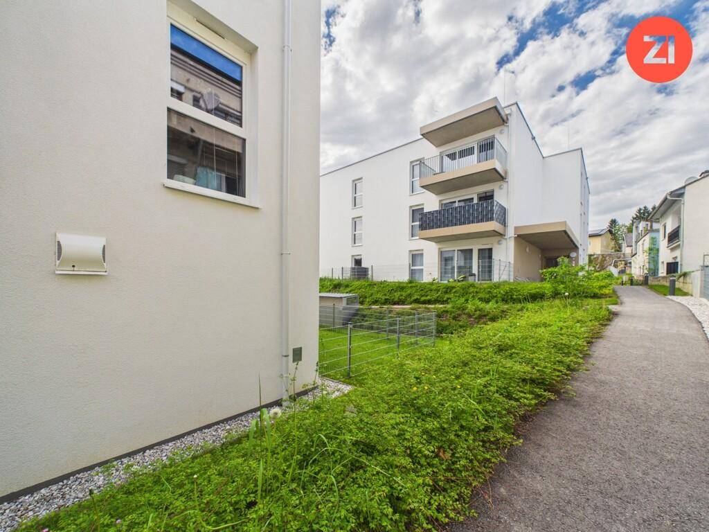 Blick entlang eines Gehwegs mit Hecke auf ein modernes Wohngebäude mit weißer Fassade.