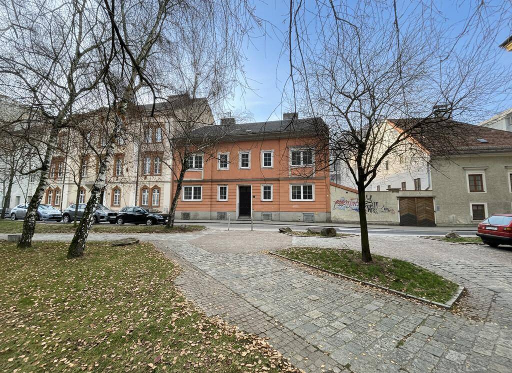 Historisches Gebäude mit orangefarbener Fassade, umgeben von Bäumen und einem gepflasterten Bereich.