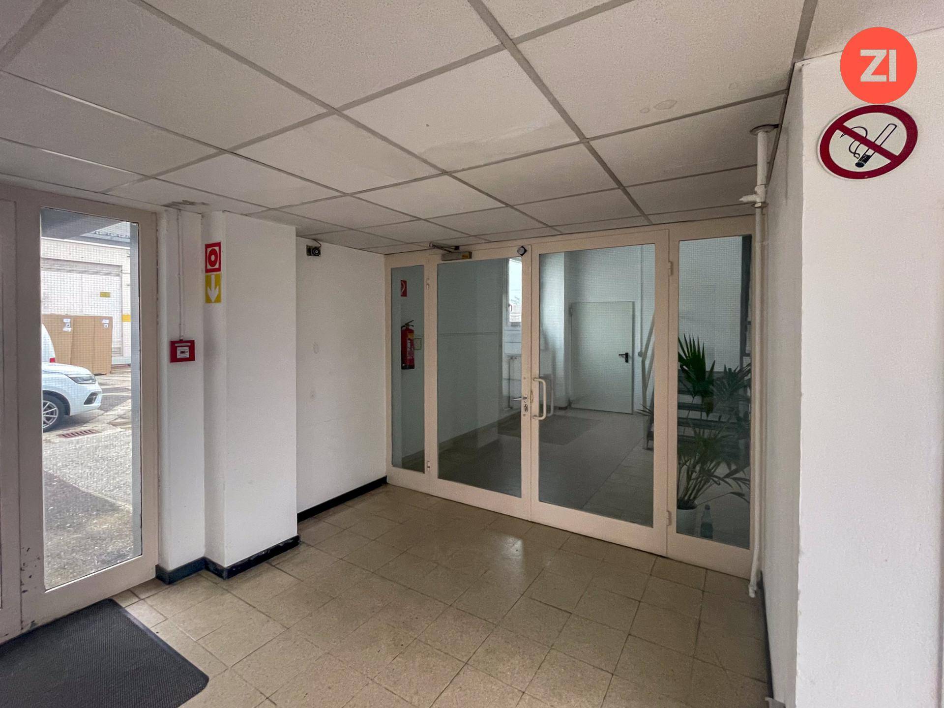 Heller Eingangsbereich mit Fliesenboden, Glastüren und einem Hinweis auf Rauchverbot an der Wand.