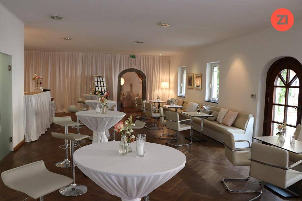 Gemütlicher Veranstaltungsraum mit Sitzgelegenheiten, Stehtischen und eleganten Vorhängen, perfekt für gesellschaftliche Anlässe.