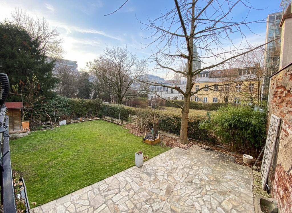Gepflegter Garten mit Rasenfläche und einer Steinterrasse, umgeben von Bäumen und Sträuchern.