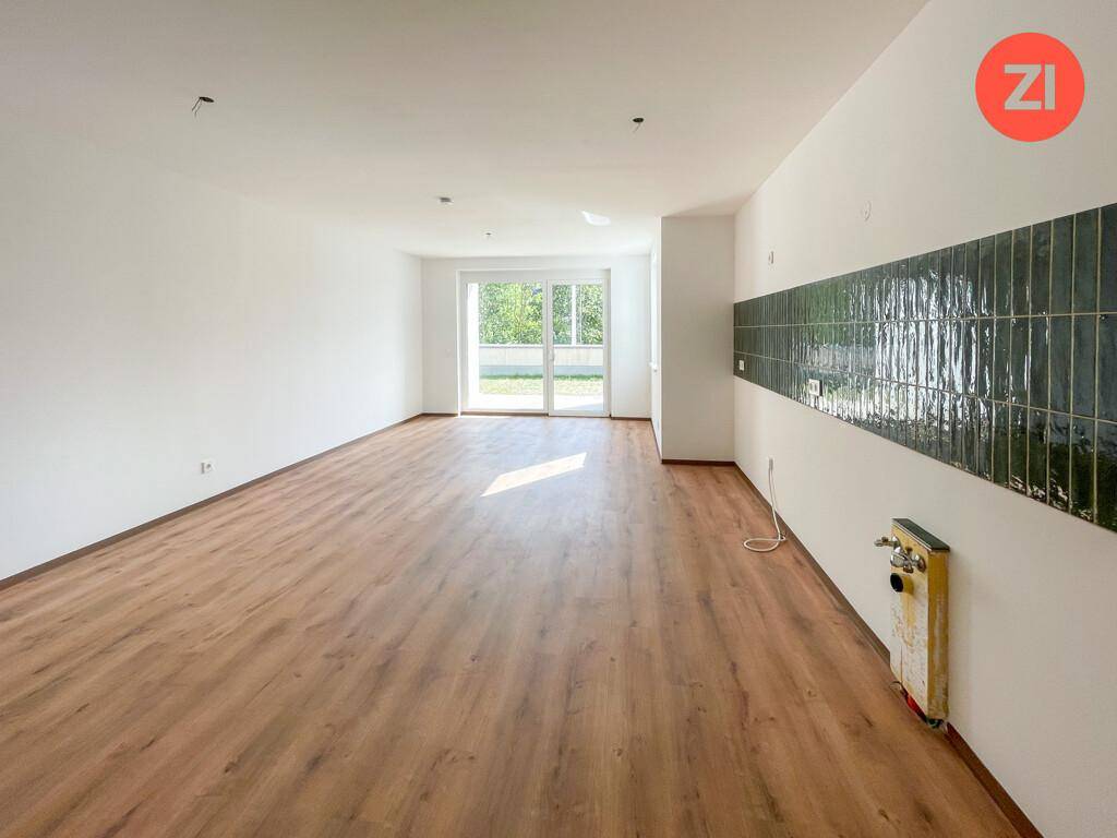 Geräumiger Wohnbereich mit Holzboden, großen Fenstern und vorbereiteter Küchenwand.