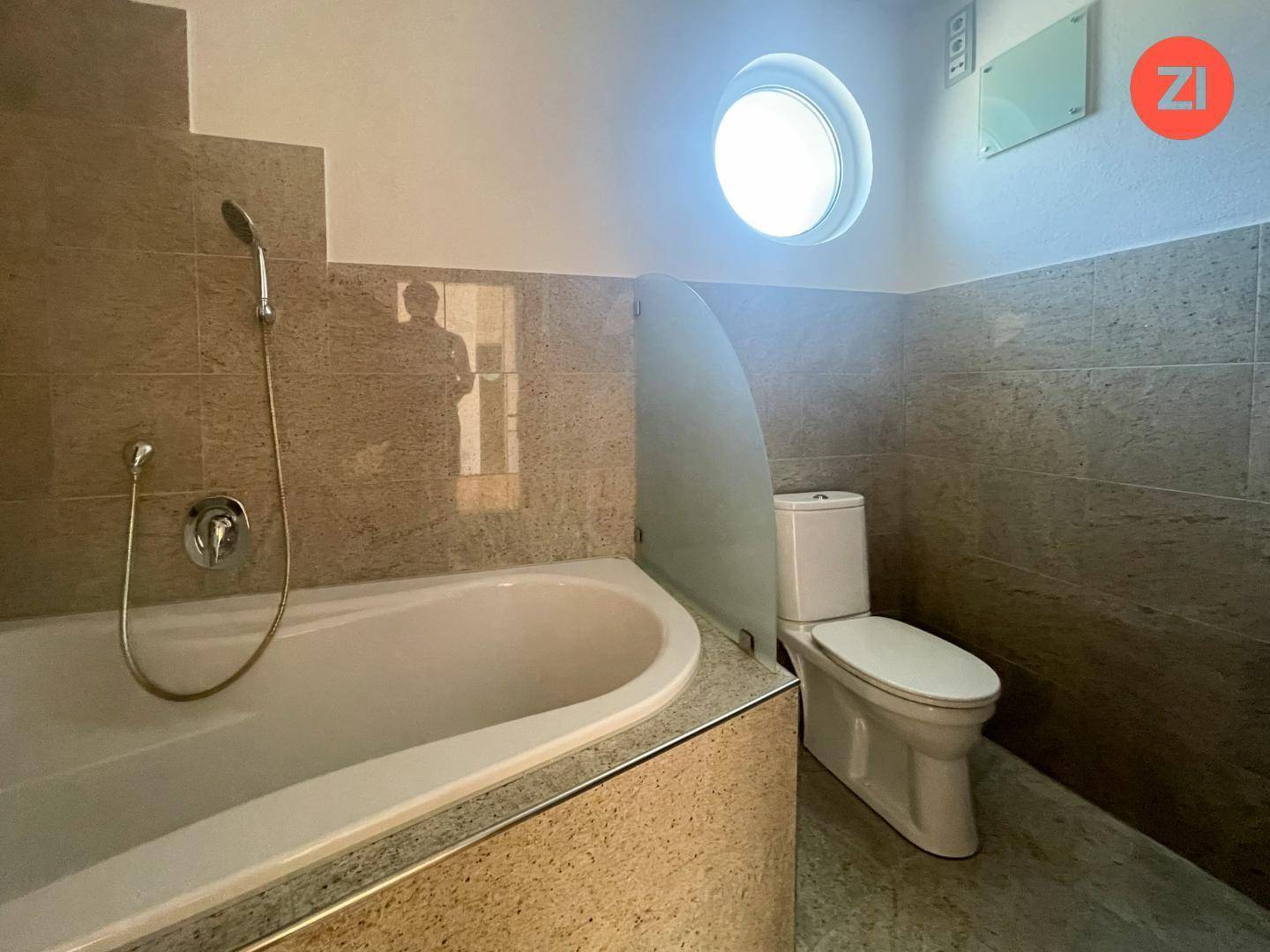 Modernes Badezimmer mit eingebauter Badewanne, Dusche und WC, ergänzt durch ein rundes Fenster.