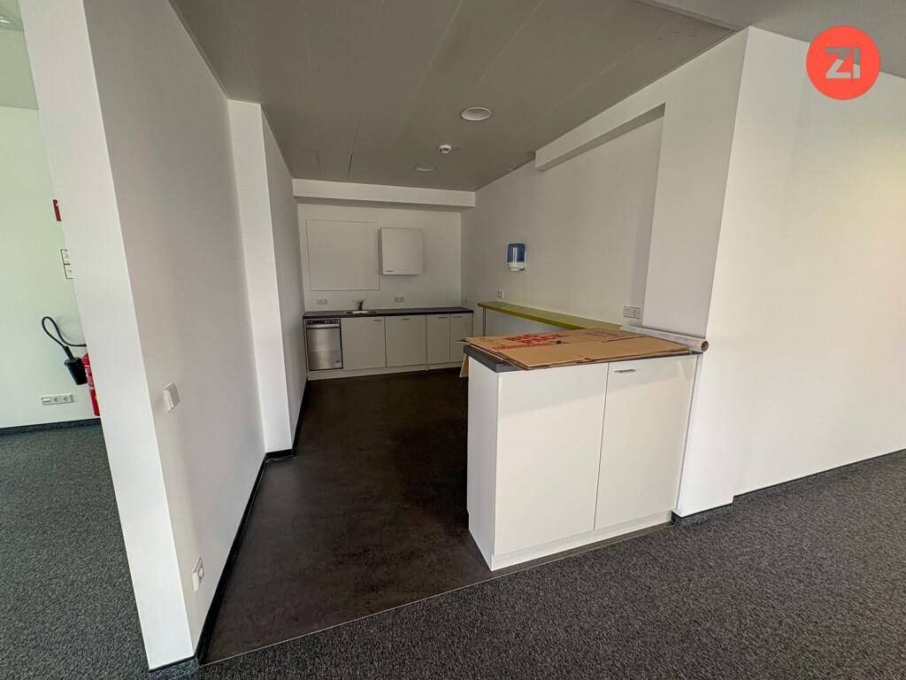 Kleine Küchenzeile mit weißen Schränken, Spüle und dunklem Boden in einem Bürobereich.