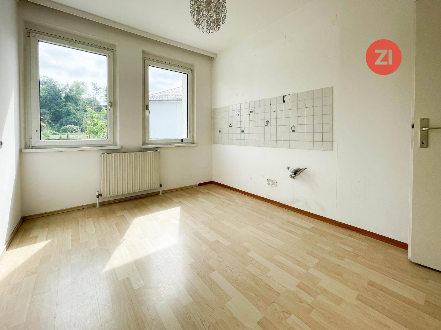 Leerer Raum, vorbereitet für eine Küche, mit Holzboden und zwei Fenstern, bereit zur individuellen Gestaltung.