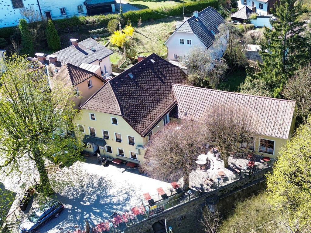 Luftaufnahme eines Gebäudes mit großem Außenbereich, möbliert mit Tischen und Stühlen, umgeben von Bäumen.