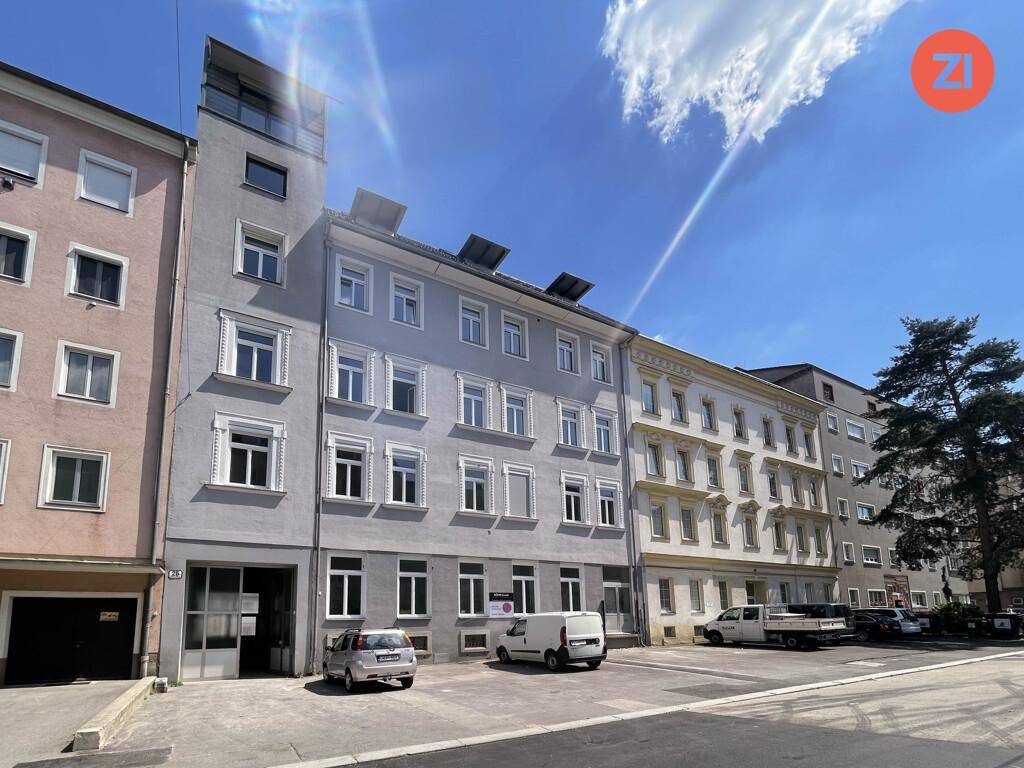 Mehrstöckiges Gebäude mit grauer Fassade, vielen Fenstern und geparkten Autos davor an einem sonnigen Tag.