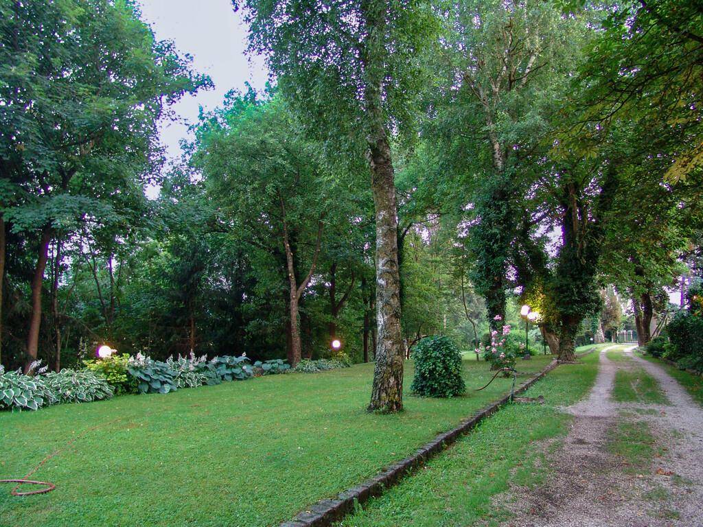 Langer Kiesweg durch einen üppigen Garten mit hohen Bäumen, Rasenflächen und dekorativer Beleuchtung.