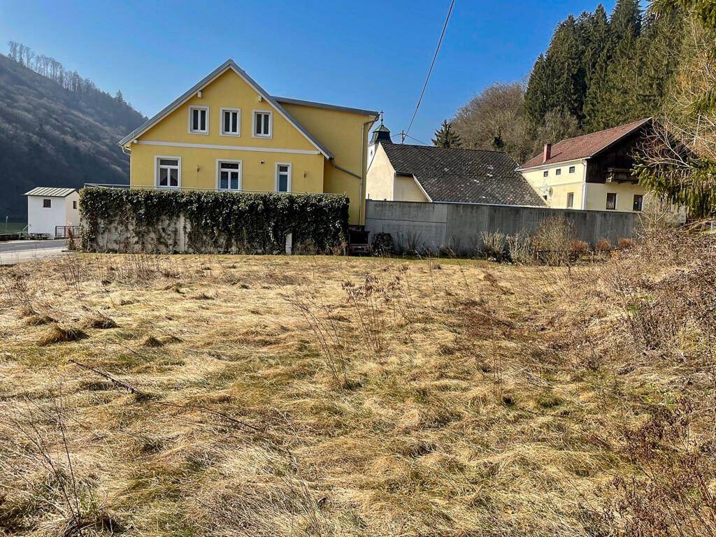 Gelbes Wohnhaus mit Hecke im Vordergrund, umgeben von grünen Hügeln und ländlicher Umgebung.
