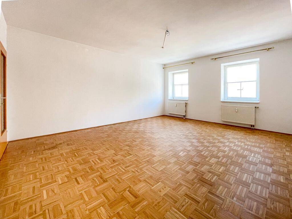 Leeres Wohnzimmer mit Parkettboden und zwei Fenstern, die viel Tageslicht hereinlassen.