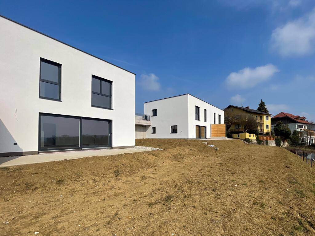 Zwei moderne Neubauhäuser mit weißer Fassade und großen Glasfronten in Hanglage.