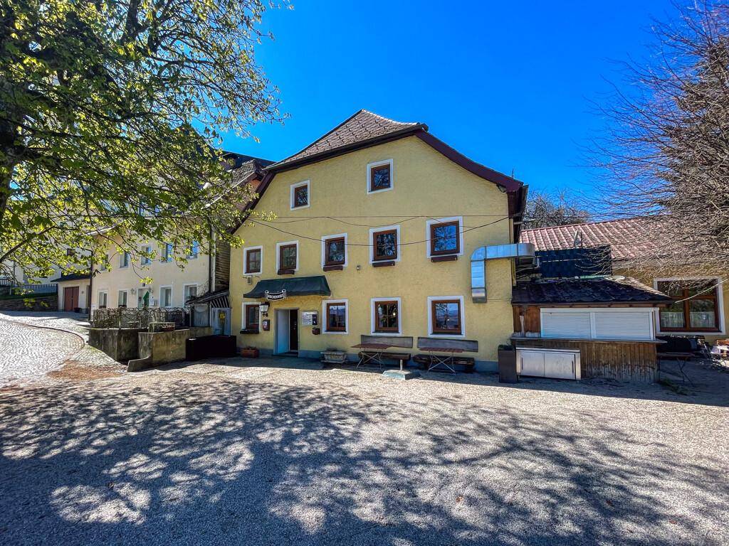 Gelbes Gebäude mit traditioneller Architektur, umgeben von Bäumen und einem gepflasterten Vorplatz unter blauem Himmel.