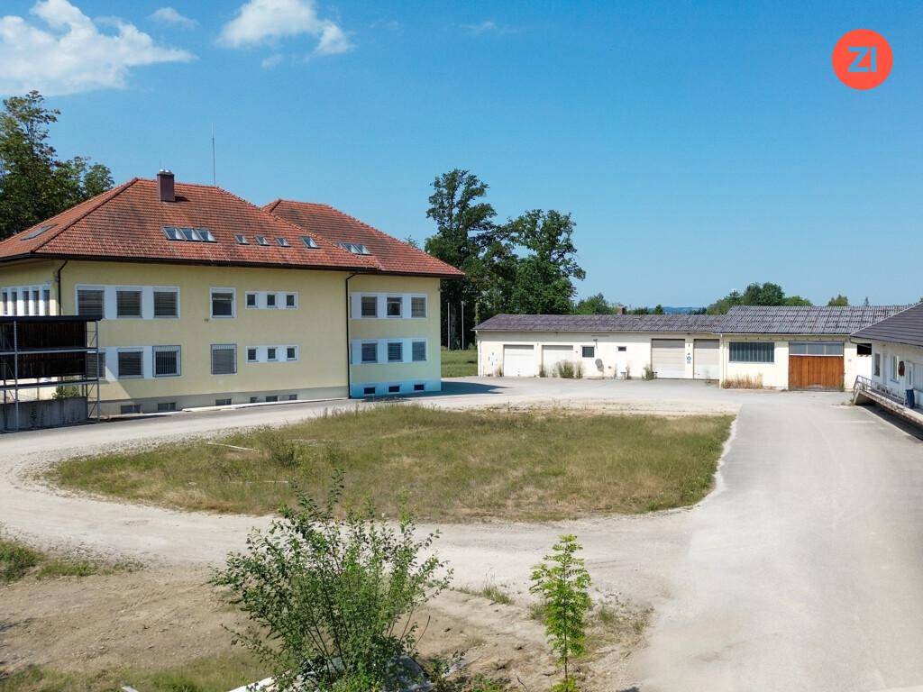 Ansicht des Hauptgebäudes und angrenzender Nebengebäude, umgeben von einer weitläufigen, unbefestigten Fläche.