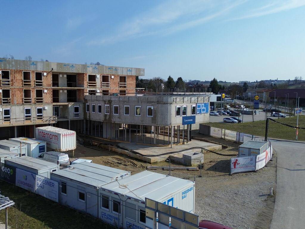 Baustelle eines Neubaus mit Rohbau, Baucontainern und Blick auf einen Supermarktparkplatz im Hintergrund.