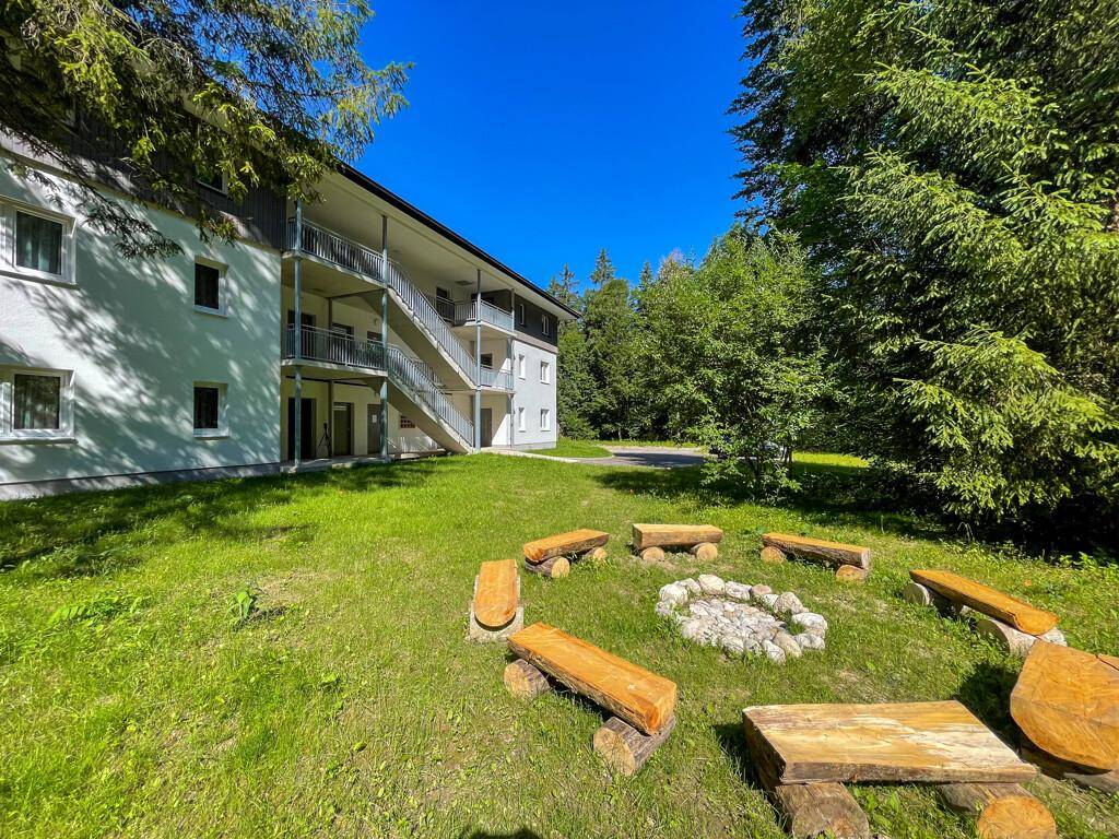 Gemütlicher Feuerstellenbereich im Garten mit Holzsitzen, umgeben von üppigem Grün und Bäumen.
