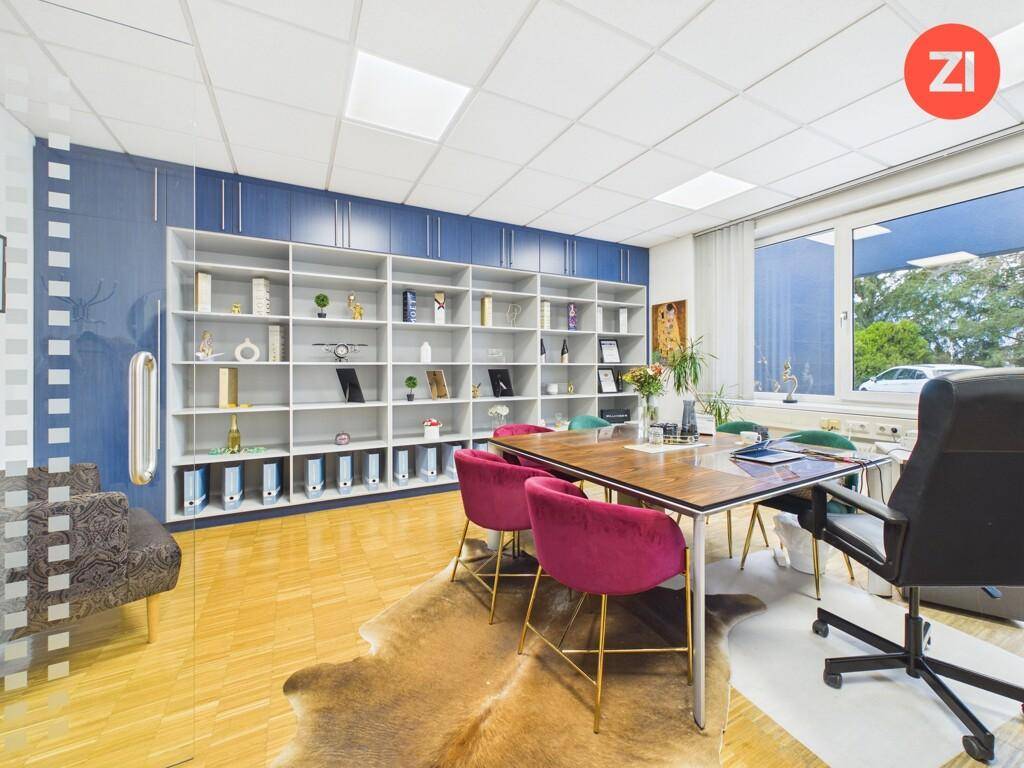 Geräumiges Büro mit Holzboden, großem Tisch, Bücherregal und Blick nach draußen durch große Fenster.