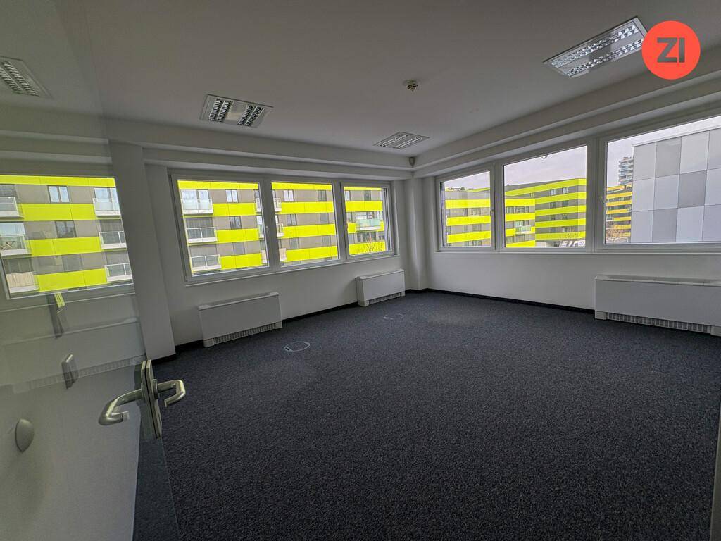 Helles Büro mit Teppichboden und großen Fenstern, die einen Blick auf ein modernes Gebäude bieten.