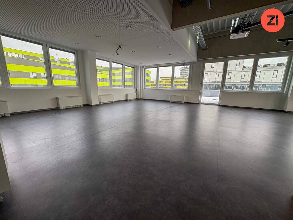 Großer, heller Büroraum mit dunklem Bodenbelag und vielen Fenstern, die viel Tageslicht hereinlassen.