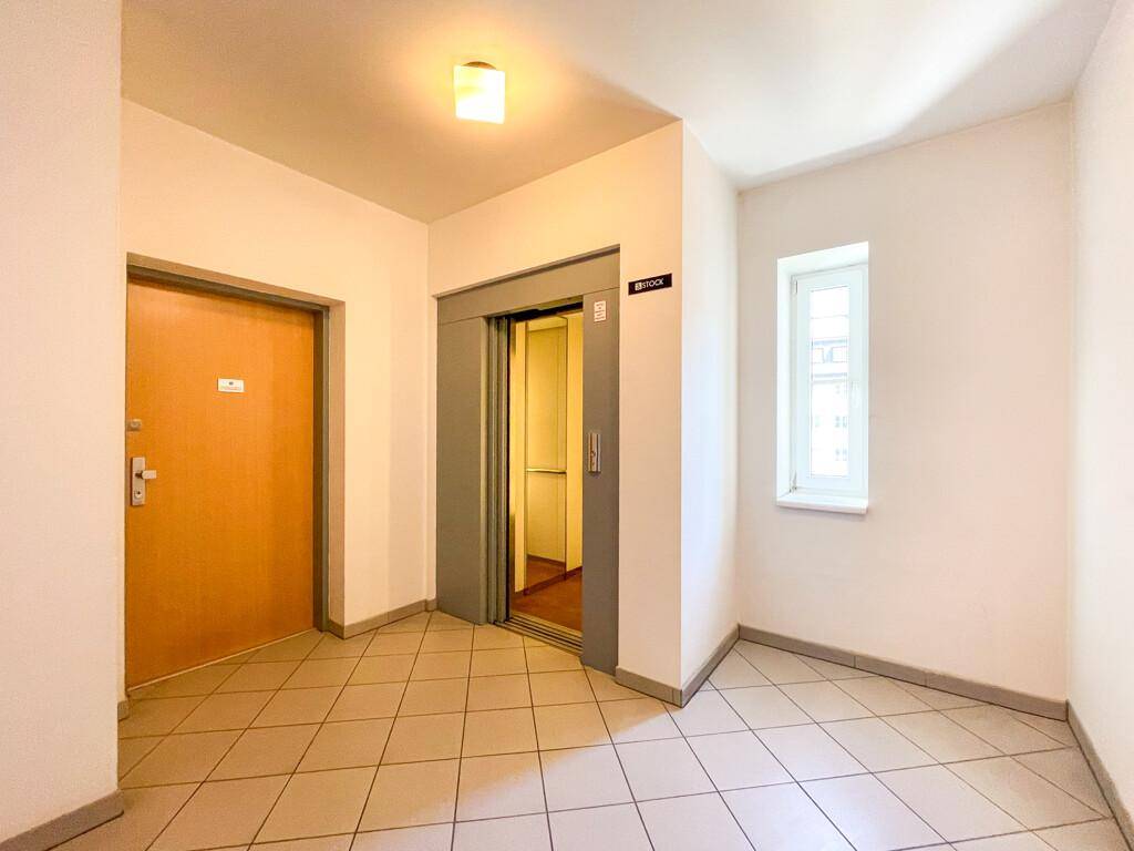 Heller Flur mit Fliesenboden, Holztür und einem Aufzug, der die Etagen verbindet.