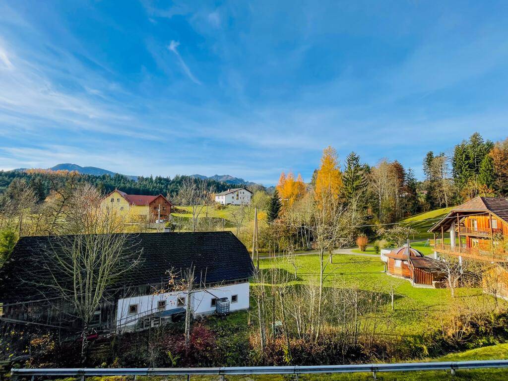 Idyllische Landschaft mit traditionellen Häusern, grünen Wiesen und herbstlichen Bäumen unter blauem Himmel.