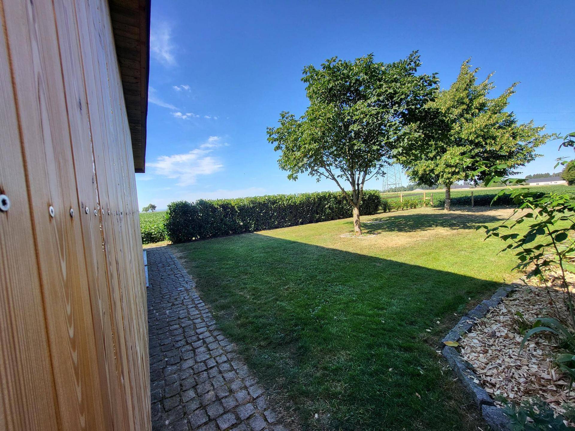 Seitlicher Gartenbereich mit Holzgebäude, gepflastertem Weg und Blick auf die umliegende Landschaft.