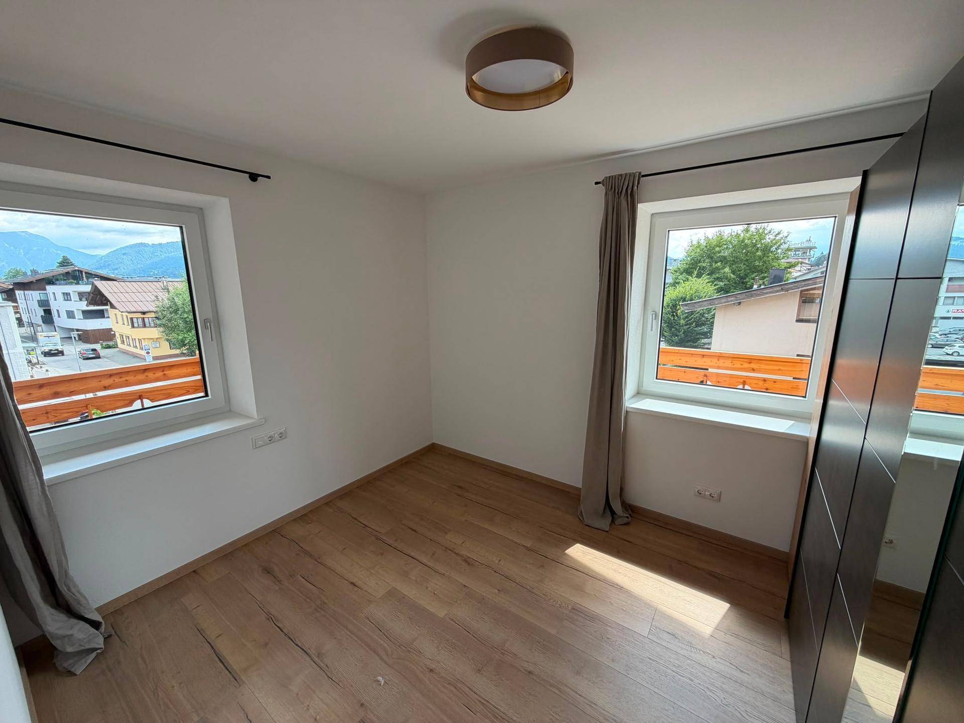 Helles Schlafzimmer mit Holzboden, zwei Fenstern und Blick ins Grüne, ausgestattet mit einem Kleiderschrank.