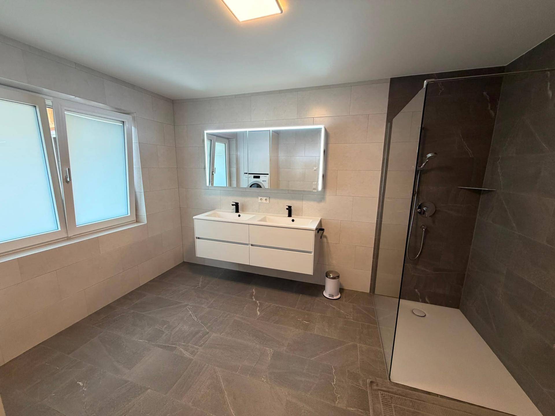 Modernes Badezimmer mit Doppelwaschbecken, großer Spiegelleuchte und ebenerdiger Dusche.
