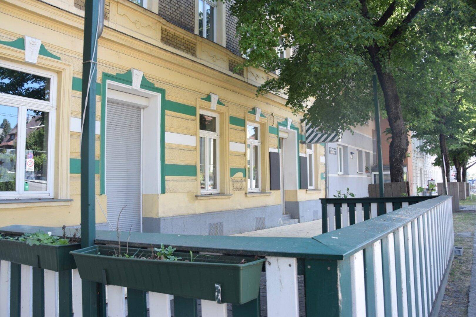 Historisches Gebäude mit gelb-grüner Fassade und einem kleinen, eingezäunten Bereich am Gehweg.