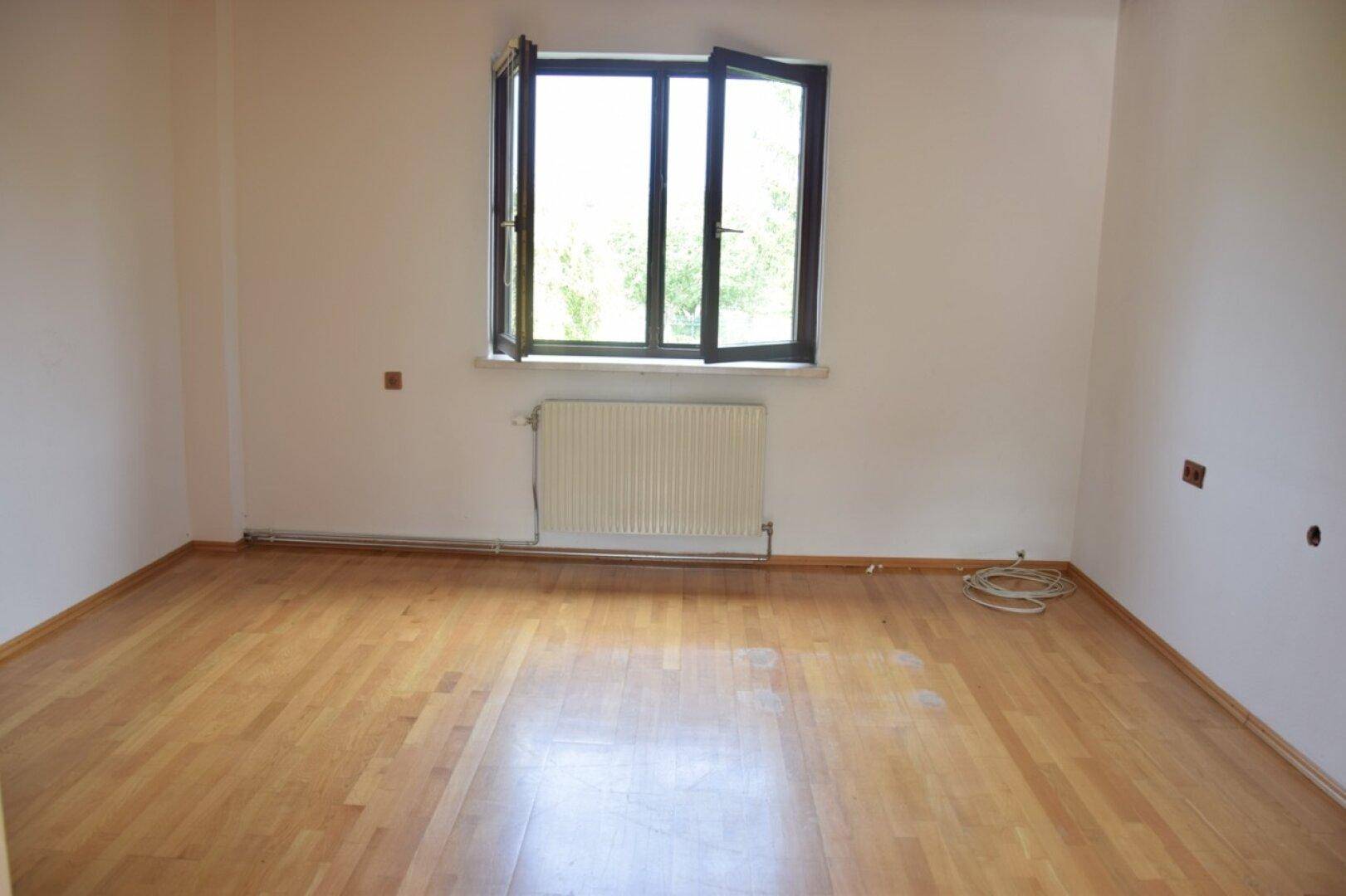 Leeres Zimmer mit hellem Holzboden, weißen Wänden und einem Fenster, bereit zur individuellen Gestaltung.