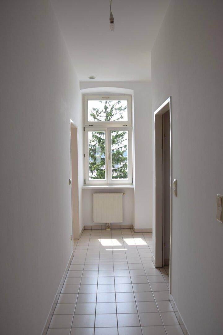 Langer Flur mit Fliesenboden und einem Fenster am Ende, das Tageslicht hereinlässt.