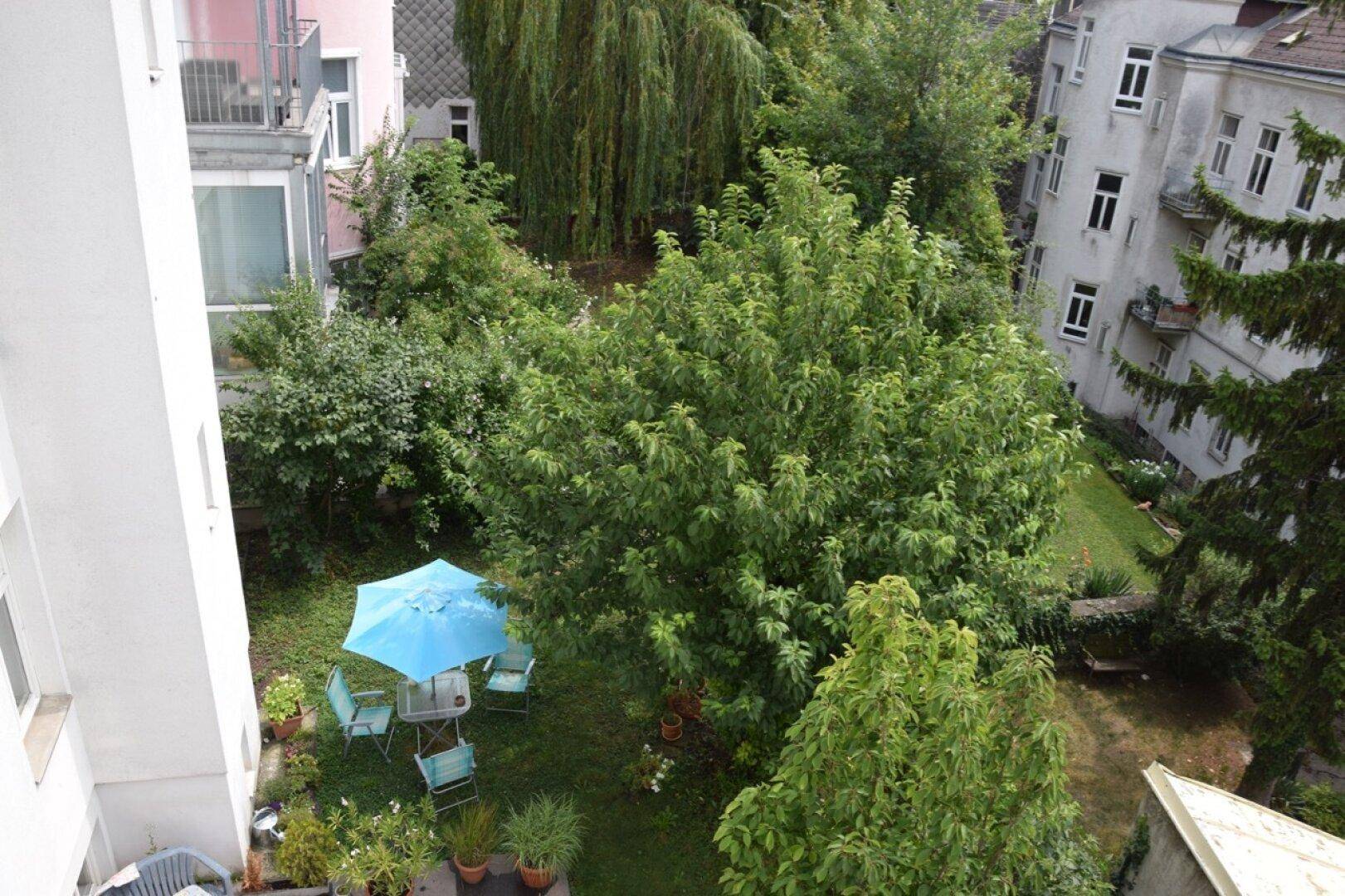 Grüner Gartenbereich mit Sitzgelegenheiten unter einem blauen Sonnenschirm, umgeben von Bäumen.