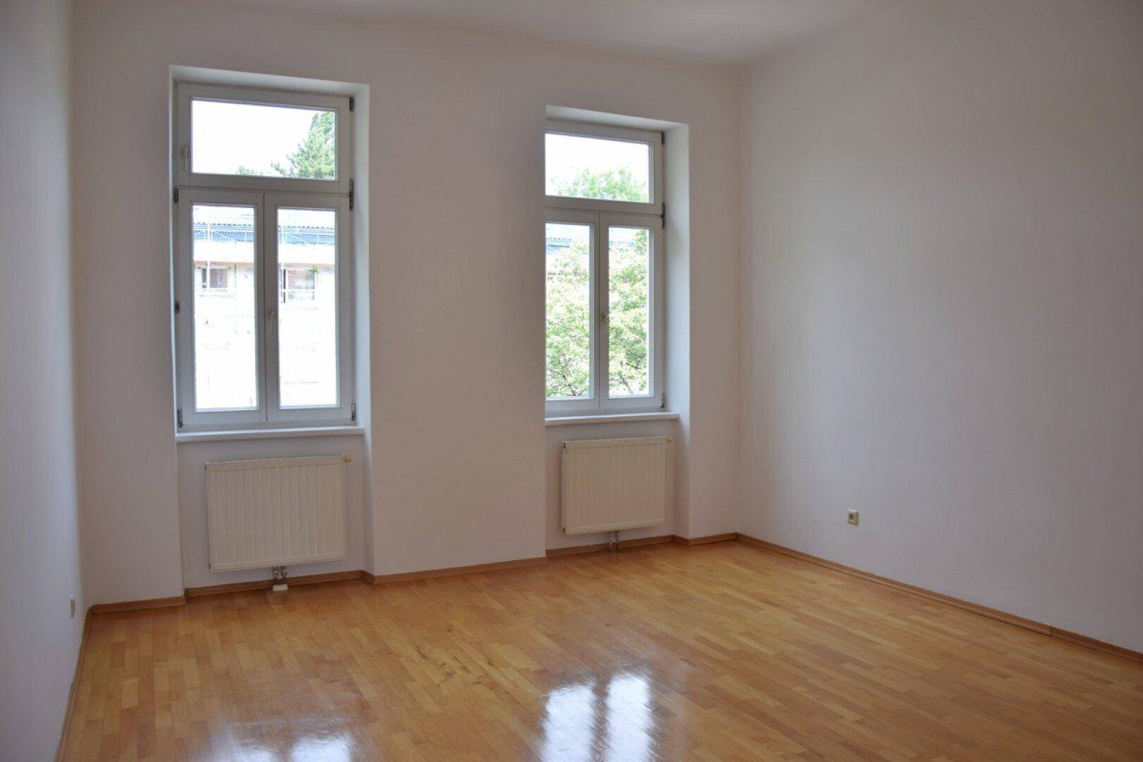 Geräumiges Zimmer mit zwei großen Fenstern und glänzendem Parkettboden.