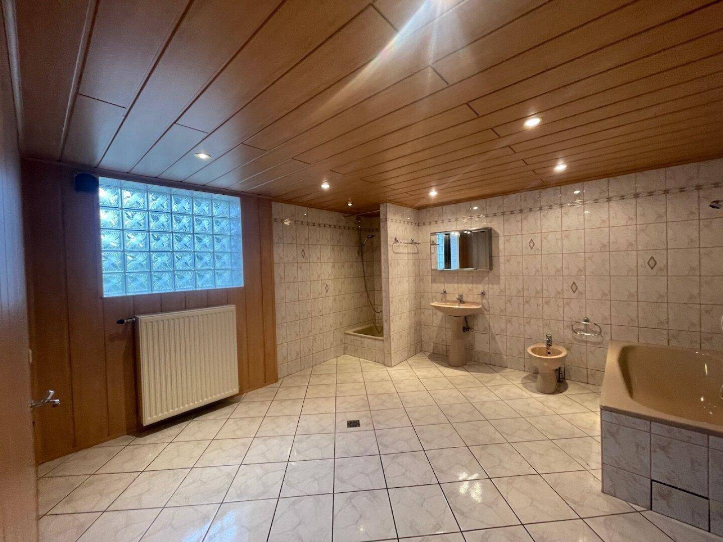 Großes Badezimmer mit weißen Fliesen, Badewanne, Bidet und Glasbausteinfenster für natürliches Licht.