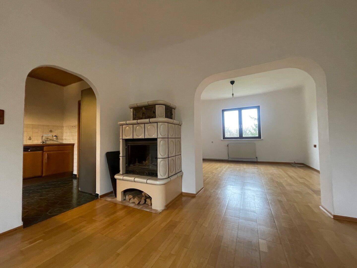 Heller Wohnbereich mit traditionellem Kachelofen und Holzboden, offen zu Küche und weiterem Raum.