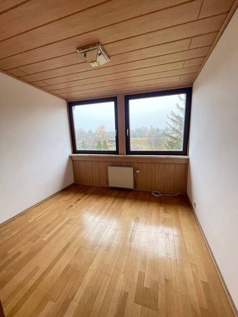 Gemütliches Zimmer mit Holzboden und Holzdecke, zwei Fenstern und einem Heizkörper.