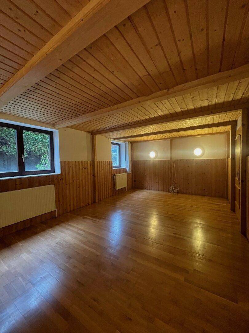Geräumiger Raum mit Holzboden, Holzvertäfelung und sichtbaren Deckenbalken, hell durch zwei Fenster.