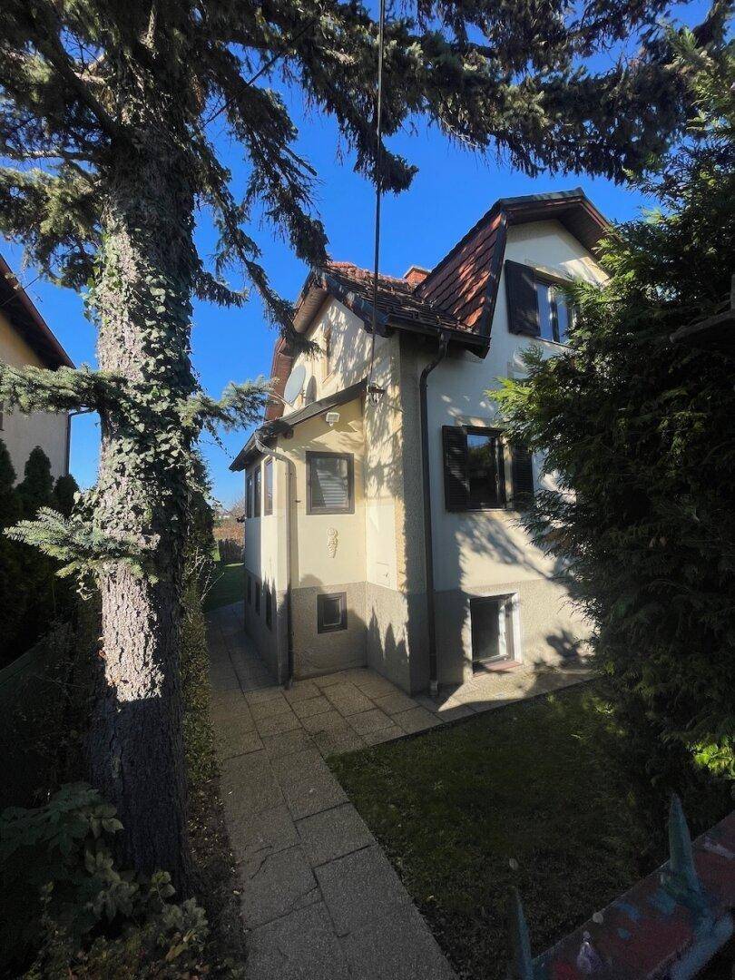 Seitenansicht des Hauses mit gepflastertem Weg und umgebenden Bäumen unter blauem Himmel.