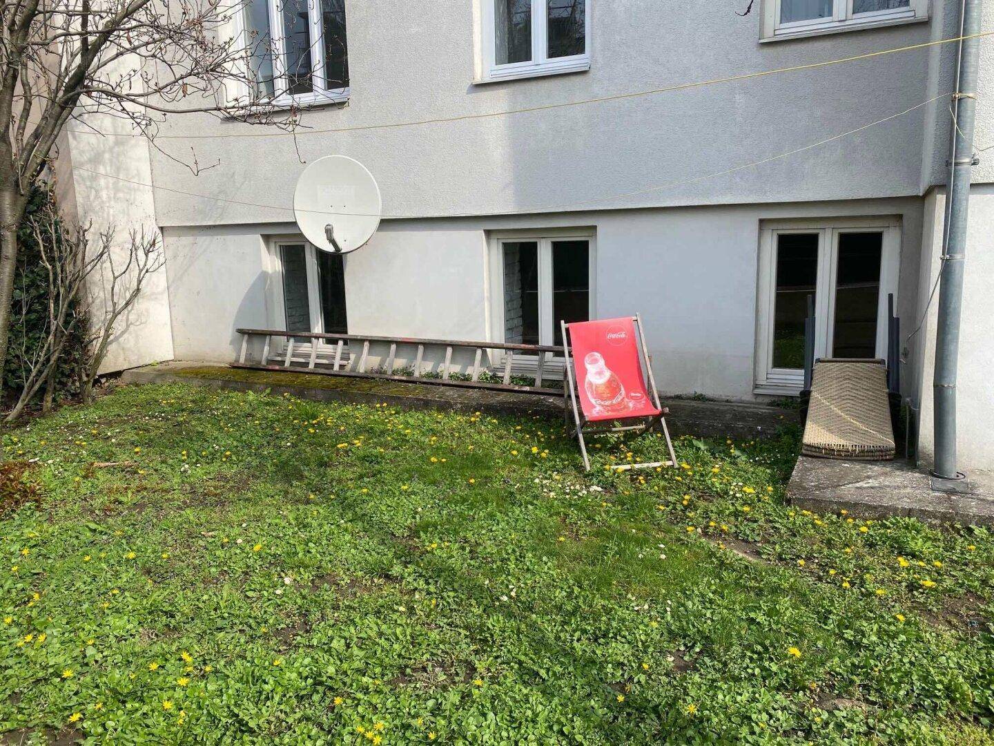 Kleiner Gartenbereich mit Rasenfläche, einer Leiter und einem Liegestuhl vor der Gebäudefassade.