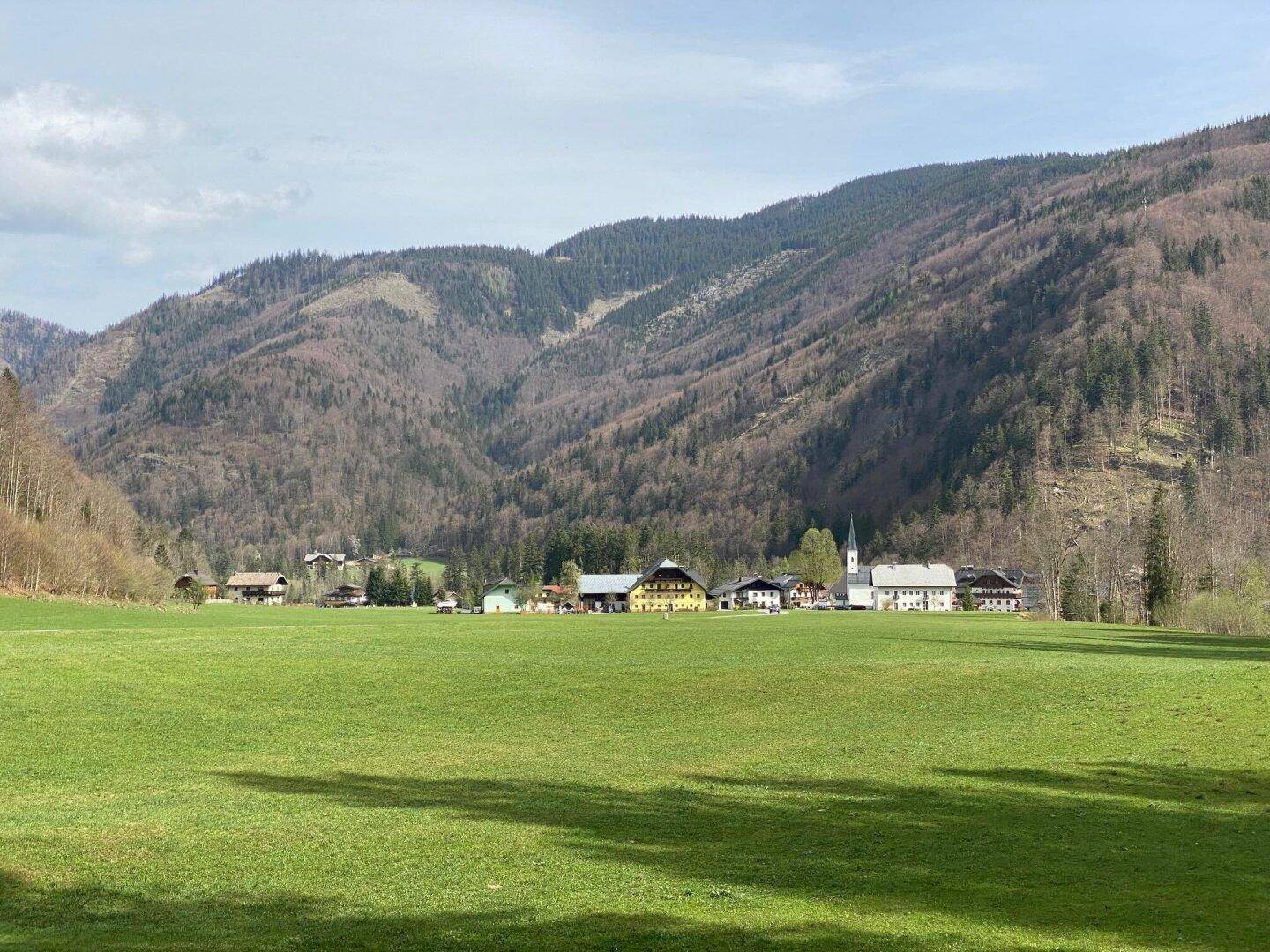 Panoramablick auf ein idyllisches Dorf in einem Tal, umgeben von grünen Wiesen und bewaldeten Bergen.