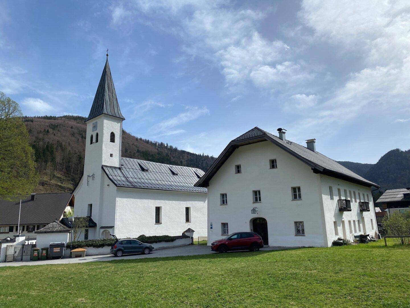 Historische Kirche und traditionelles Gebäude in einer ländlichen Berglandschaft unter blauem Himmel.