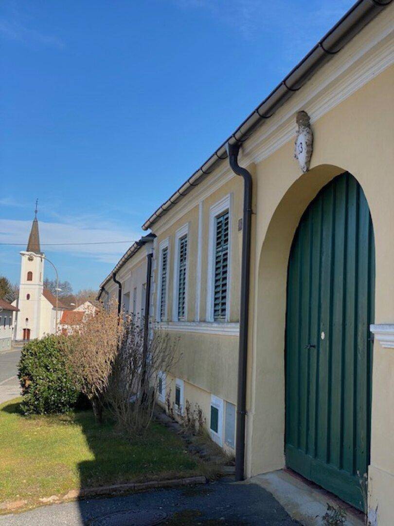 Seitenansicht eines traditionellen Hauses mit großer grüner Holztür und Kirchturm im Hintergrund.