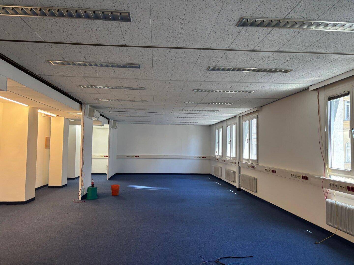 Großes, leeres Büro mit blauem Teppichboden, vielen Fenstern und abgehängter Decke.