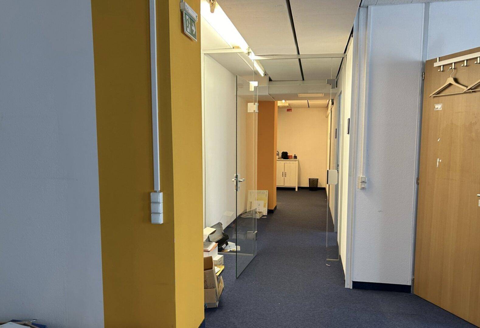 Bürogang mit blauem Teppichboden, Glaswänden und gelben Akzenten, führt zu weiteren Büroräumen.