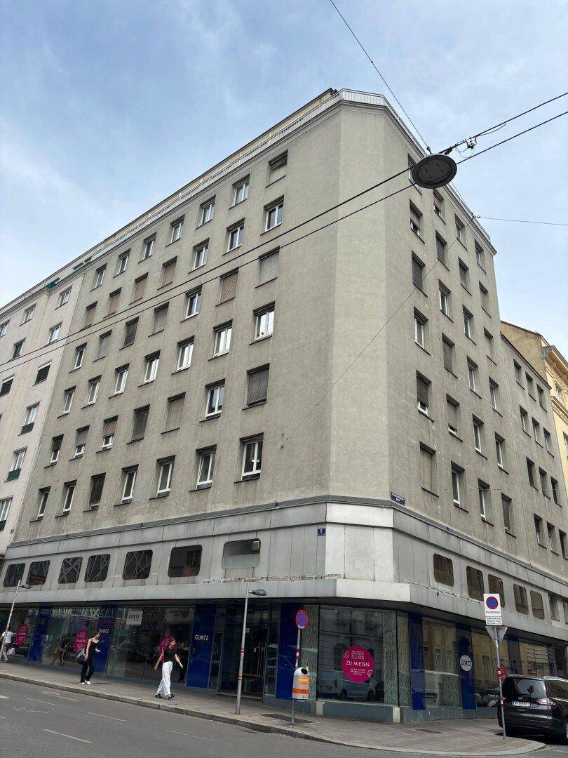 Mehrstöckiges Gebäude mit vielen Fenstern und Geschäftsflächen im Erdgeschoss an einer Straße.
