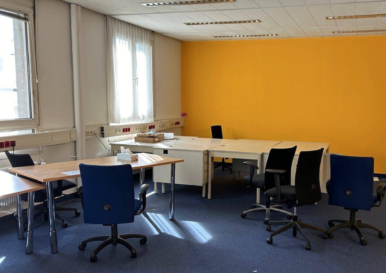 Möblierter Büroraum mit Schreibtischen, Bürostühlen, blauem Teppich und einer gelben Akzentwand.