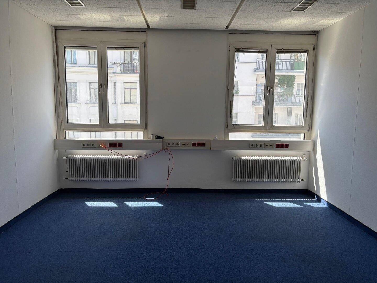Leerer Büroraum mit zwei Fenstern, Heizkörpern darunter und blauem Teppichboden.
