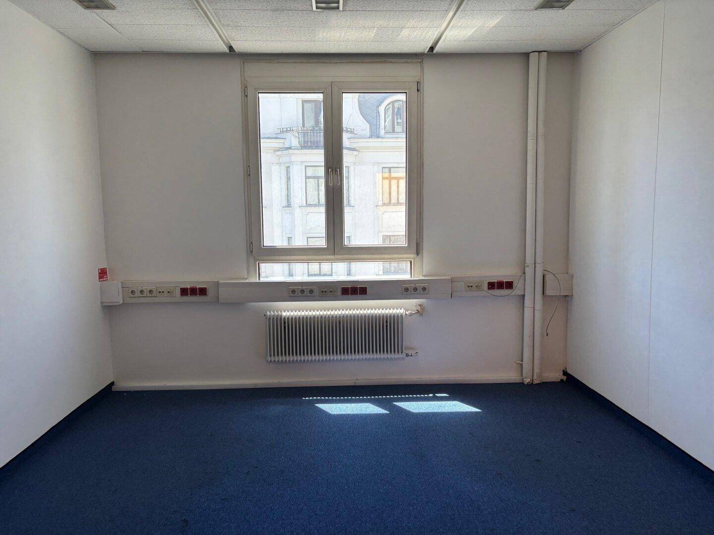 Leerer Büroraum mit einem Fenster, Heizkörper und blauem Teppichboden.