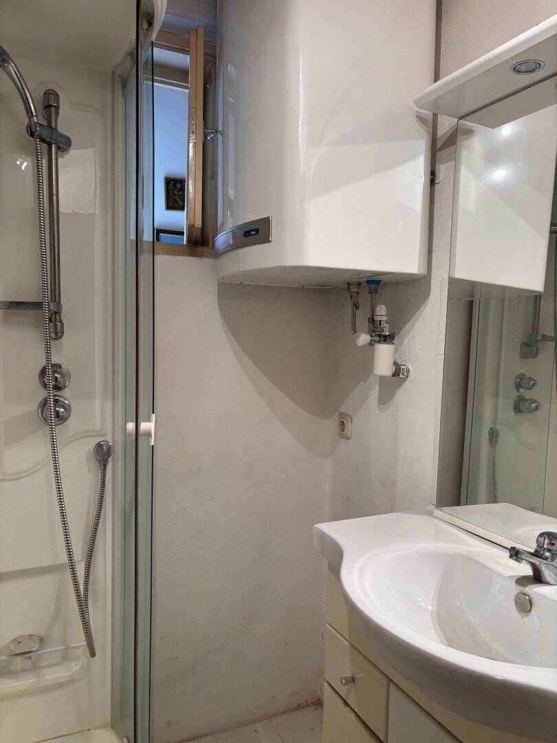 Kleines Badezimmer mit Duschkabine, Waschbecken und Warmwasserboiler.