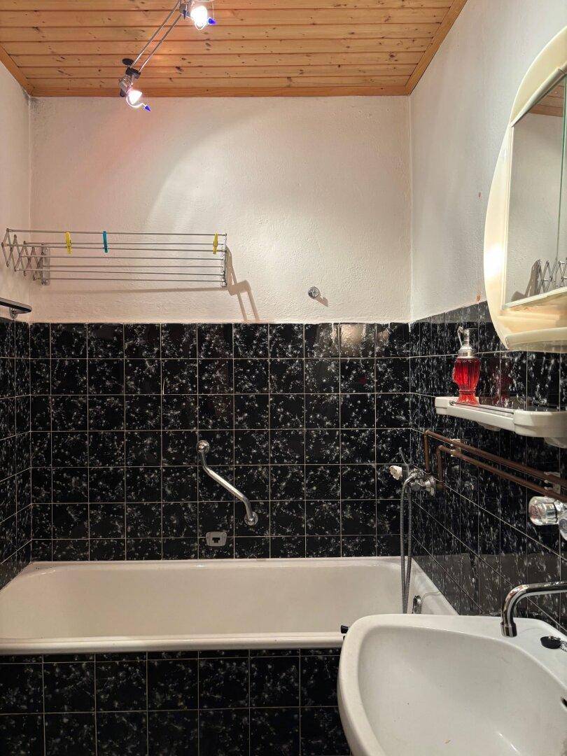 Badezimmer mit Badewanne, Waschbecken und dunklen Fliesen im 80er-Jahre-Stil.